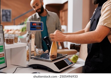 At checkout counter caucasian vendor places brown paper bag on weighing scale to check value for male customer. Arab man waiting at cashier desk while shopkeeper measures his eco friendly parcel. - Powered by Shutterstock