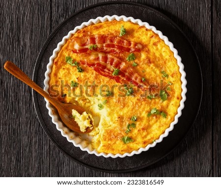 Cheddar Cheese Grits Casserole with bacon slices in baking dish with spoon on dark wooden table, horizontal view from above, flat lay, close-up