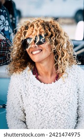 Cheeful and beautiful curly blonde caucasian adult young woman smiling and laughing with sunglasses and blue vintage van in background - concept of happy people