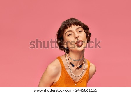 cheeky carefree girl in 20s with short brunette hair sticking tongue out on pink background