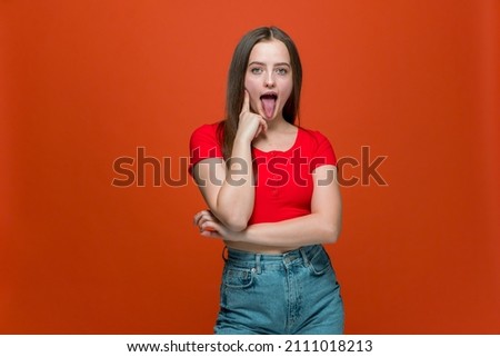 Cheeky modern young girl showing tongue, teasing, making faces, fooling, grimacing on orange studio background