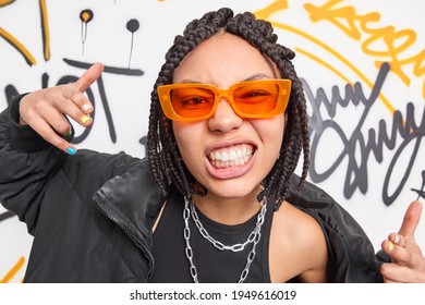 Cheeky woman with dreadlocks clenches teeth makes yo gesture feels cool belongs to street gang wears trendy orange sunglasses fashionable jacket poses against graffiti wall. Hooliganism concept - Powered by Shutterstock