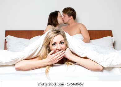 Cheeky young woman in a threesome or the cheating partner in an affair peeking out of the bottom of the bedclothes with a saucy expression as a young couple at the top of the bed share a loving kiss