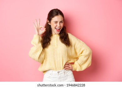 Cheeky young woman winking, showing okay sign, give her approval, like and approve good thing. Girl make OK gesture to give permission, say yes, standing over pink background