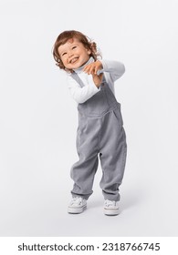 A cheerful 2-year-old toddler with curly hair is indulging, laughing, smiling, holding his hands to himself and bending over in a gray jumpsuit and a white turtleneck