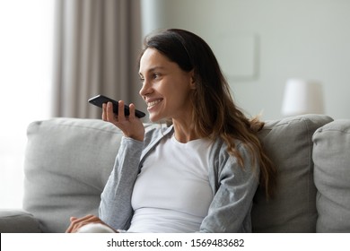 Cheerful 30s woman sitting on sofa holding smartphone near mouth talks with friend on loudspeaker through built-in mic. Usage of voice messaging, modern ai tech, intelligent virtual assistant concept