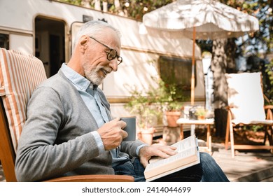 Cheerful active caucasian senior old man grandfather reading book drinking coffee while on a trip voyage by trailer camper van motor wheel home