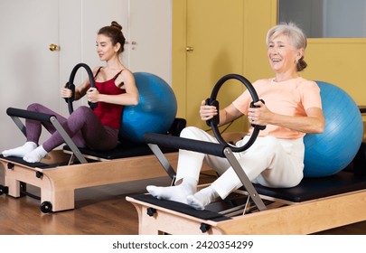 Cheerful active elderly woman exercising with flex ring and fitball on reformer in pilates studio during group workout - Powered by Shutterstock