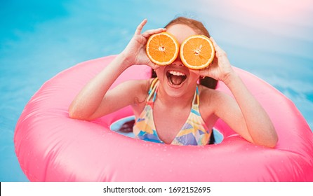 Cheerful Active Little Girl In Swimsuit Covering Eyes With Sliced Orange Fruit While Having Fun In Pool With Pink Inflatable Ring