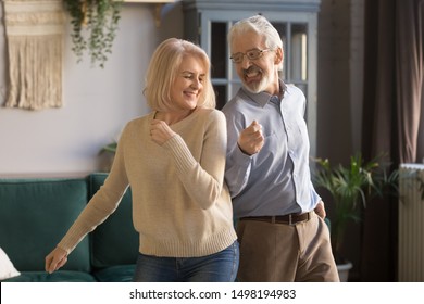 Cheerful Active Middle-aged Mature Husband And Wife Have Fun In Living Room Together Moving To Rhythm, Happy Overjoyed Senior Spouses Or Couple Dance Enjoy Leisure Weekend Activity Relaxing At Home