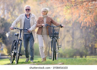 Cheerful Active Senior Couple With Bicycles Walking Through Park Together. Perfect Activities For Elderly People.