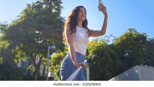 Cheerful active woman rising stairs making photo beautiful city park. Happy asian lady using modern smartphone to take picture cityscape. Attractive girl tourist enjoy photographing outdoors.