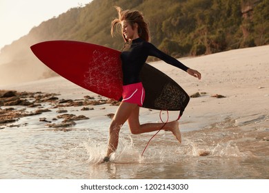 Cheerful Active Woman Runs Into Water With Happy Expression, Wants To Show Duck Dive, Being Goofy, Dressed In Special Clothes For Surfing, Beautiful Marine Scenery In Background. Leisure Concept