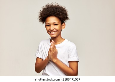 Cheerful adorable teen kid laughing with palms pressed together, dressed in white t-shirt. African american boy of 12 thanking for support smiling at camera with folded hands. Happy carefree childhood - Shutterstock ID 2188491089