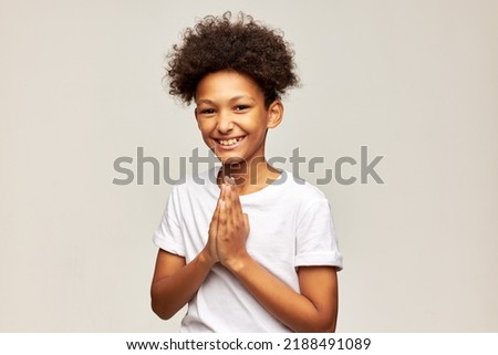 Cheerful adorable teen kid laughing with palms pressed together, dressed in white t-shirt. African american boy of 12 thanking for support smiling at camera with folded hands. Happy carefree childhood
