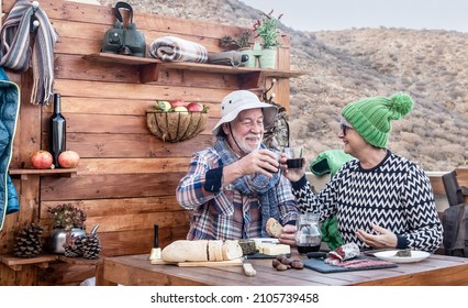 Cheerful adult aged senior couple enjoying a break with food and drink toasting with red wine. Wooden table with traditional products of the mountain. Smiling active retired elderly people in holiday
