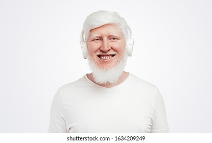 Cheerful adult albino male in white t shirt listening music with white headphones while standing against white background - Powered by Shutterstock