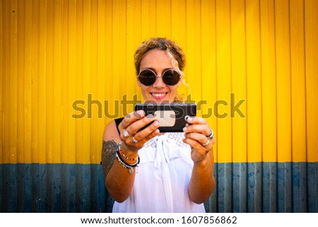 Cheerful adult beautiful woman taking selfie picture with modern phone and urban yellow and blue background - modern people enjoying social media technology  and internet connection outdoor