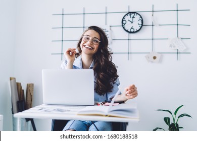 Cheerful Adult Dark Haired Woman In Casual Clothes And Glasses Looking At Camera While Sitting At Desk And Rising Hand Against White Wall With Black Decor And Clock In Light Minimalistic Apartment