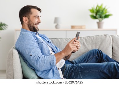 Cheerful adult european guy with beard sits on sofa chatting on phone in living room interior, side view. Great offer, advertising and video chat on device, social distance and new normal, copy space