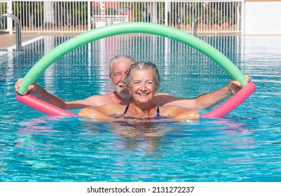 Cheerful adult happy senior couple having fun in outdoors swimming pool doing exercise with swim noodles. Smiling retired people playing together in the pool water under the sun