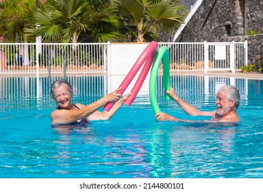 Cheerful adult happy senior couple having fun in outdoors swimming pool doing battle with swim noodles. Smiling people playing together in the pool enjoying good time and vacation