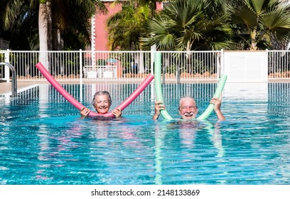 Cheerful adult happy senior couple having fun in outdoors swimming pool with swim noodles. Smiling retired people playing together in the pool water under the sun enjoying vacation