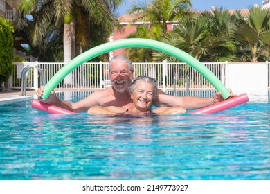 Cheerful adult happy senior couple having fun in outdoors swimming pool doing exercise with swim noodles. Smiling retired people playing together in the pool water under the sun