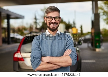 Cheerful adult male customer with beard in smart casual outfit and eyeglasses smiling happily, and looking at camera while standing near mode SUV car at gas station with crossed arms