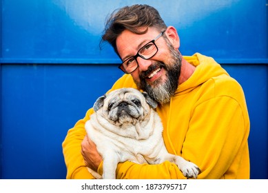 Cheerful adult man smile and hug with love his own old dog pug in a portrait with yellow and blue color - people with animals and glasses enjoy the pet therapy concept