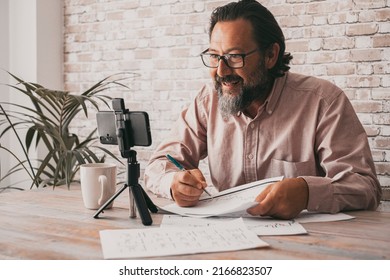 Cheerful adult man working at home office using mobile phone to film contents for the web.. Concept of video call conference and online job. Content creator male people writing and speaking happy