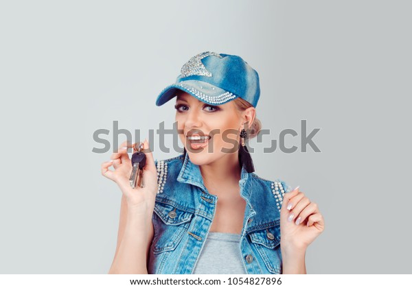Cheerful adult pretty glamour woman in denim cap\
and clothes showing automobile keys on gray background. Horizontal\
image shot in studio