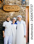 Cheerful adult smiling female workers in white uniforms embracing each other outside of cheese factory with wooden Formatgeria signboard