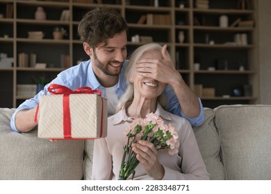 Cheerful adult son guy giving surprise gift to old senior mom, covering woman eyes with hand, holding present box, flowers bouquet, celebrating international women day, birthday, mothers day - Shutterstock ID 2283348771