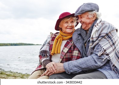 Cheerful And Affectionate Seniors Sitting By Seaside In Autumn