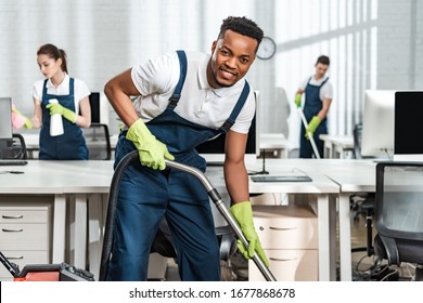 cheerful african american cleaner vacuuming floor while looking at camera