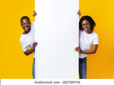 Cheerful African American Couple In Love Holding Empty Board For Advertisement Or Text Over Yellow Studio Background. Smiling Black Man And Woman Standing Next To Blank Placard For Advert