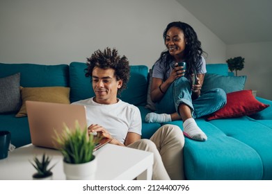 Cheerful African American Couple Spending Time In The Living Room On The Couch And Looking At Laptop Screen And Drinking Coffee While Enjoying Weekend Together.