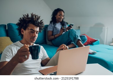 Cheerful African American Couple Spending Time In The Living Room On The Couch And Looking At Laptop Screen And Drinking Coffee While Enjoying Weekend Together.