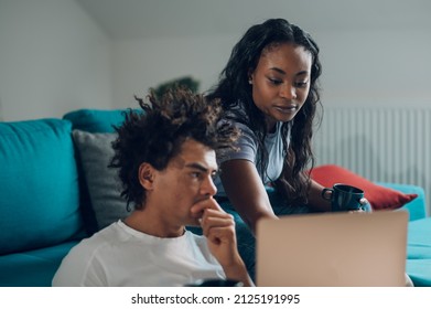 Cheerful African American Couple Spending Time In The Living Room On The Couch And Looking At Laptop Screen And Drinking Coffee While Enjoying Weekend Together.