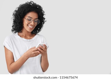 Cheerful African American with crisp hair, holds modern smart phone, happy to recieve message, dressed in casual t shirt, poses against white background with blank space for your advertisement - Powered by Shutterstock