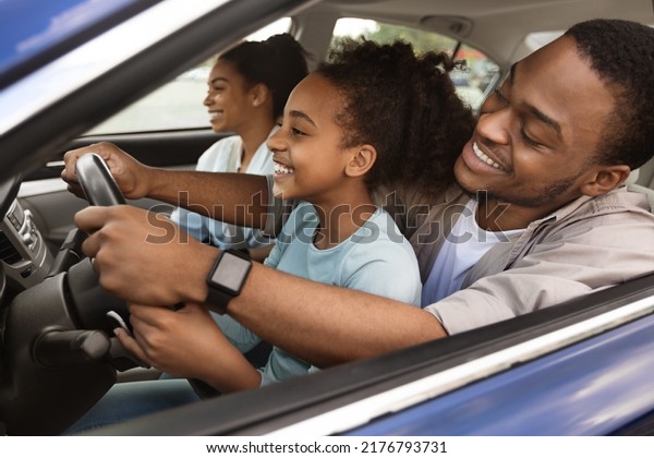 Cheerful\
African American Dad Teaching Preteen Daughter To Drive A Car. Kid\
Sitting On Father\'s Knees Driving Together And Having Fun While\
Family Traveling By Automobile. Selective\
Focus