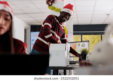 Cheerful african american employee wearing Christmas hat unpacking belongings box, decorating new desk after being hired. Businessman starting job, excited to work in festive ornate office - Powered by Shutterstock