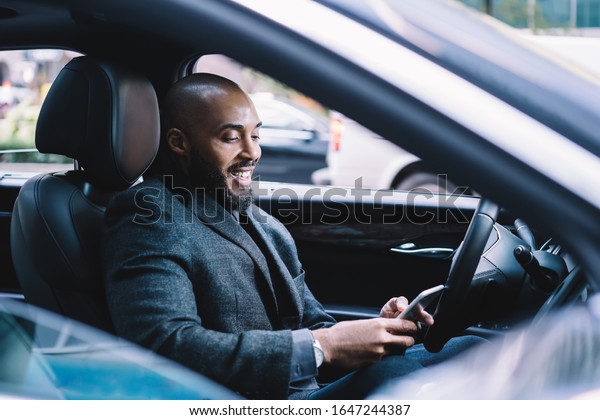 Cheerful African\
American executive man in elegant gray jacket focusing on screen\
and interacting with smartphone with pleasure while driving car\
standing in traffic jam in\
downtown