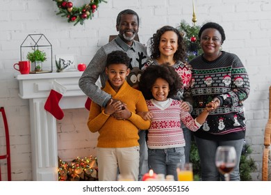 Cheerful African American Family Holding Hands And Looking At Camera Near Fireplace With Christmas Decoration