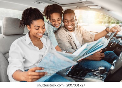 Cheerful African American Family Looking At Road Map Sitting In Car. Parents And Daughter Choosing Destination For Summer Road Trip Together. Local Tourism Concept. Selective Focus