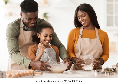 Cheerful African American Family Making Dough Baking Cake Together Mixing Ingredients In A Bowl In Modern Kitchen At Home. Parents And Daughter Making Cookies On Weekend