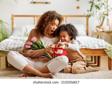 Cheerful african american family mom opens a gift box with her son and holding bouquet  of flowers while resting on floor by bed during holiday celebration mothers day at home - Shutterstock ID 2133356817