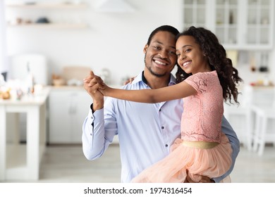 Cheerful African American Father Dancing With His Little Cute Daughter In Beautiful Dress, Holding Her On Hands, Kitchen Interior, Copy Space. Happy Black Family Enjoying Time Together At Home