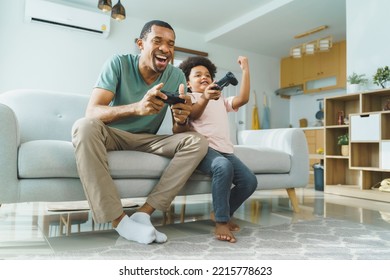 Cheerful African American Father and his Little son in Afro hair sitting on sofa using joysticks or game controllers playing console video games at home - Powered by Shutterstock